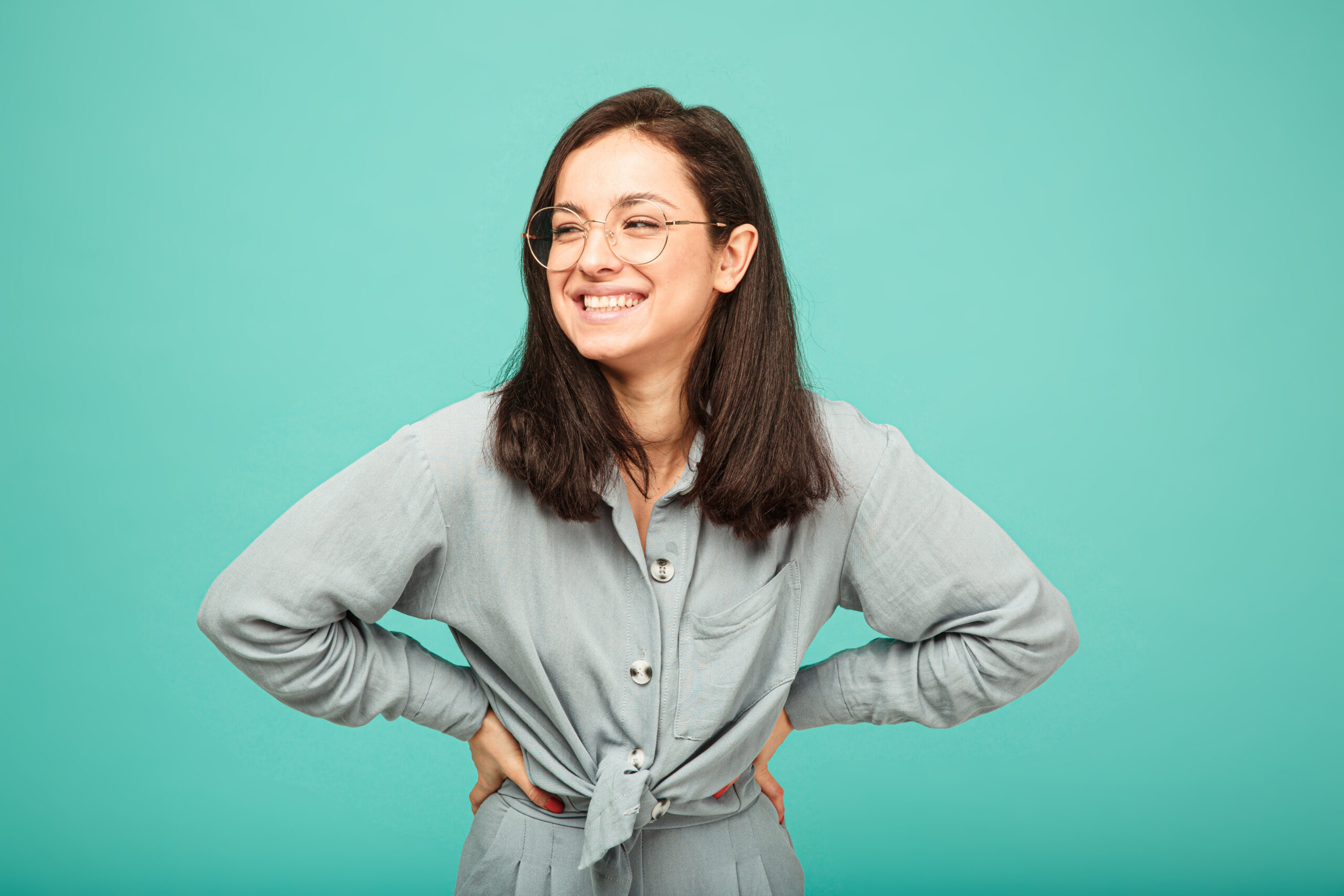 Photo of cute female in glasses smiles nicely. Wears grey shirt, isolated turquoise color background.